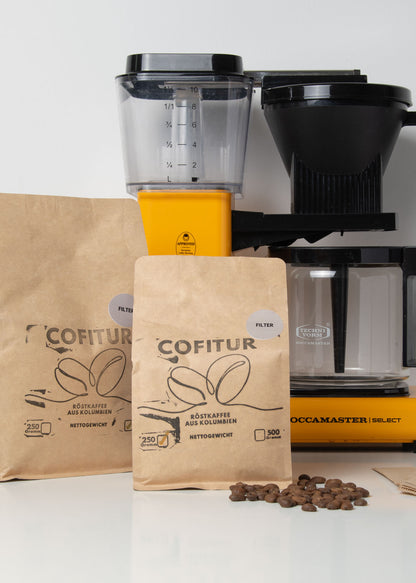 Abgepackter Cofitur Filterkaffee in 250g- und 500g-Papiertüten – im Hintergrund eine Moccamaster-Kaffeemaschine auf einer weißen Fläche