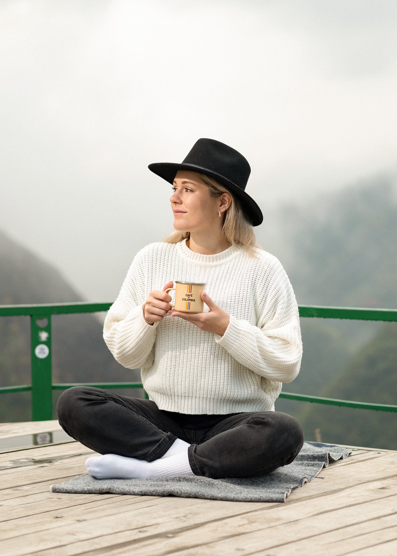 Vera Richartz mit Kaffeetasse und Hut sitzt im Schneidersitz vor dem Nebelwald in Huila – achtsamer Moment auf der Cofitur-Finca mit Blick auf die Kaffeepflanzen am Hang